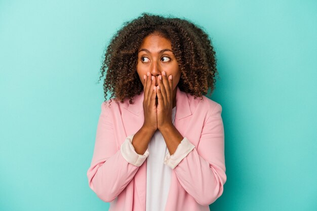 Jeune femme afro-américaine aux cheveux bouclés isolée sur fond bleu réfléchie à la recherche d'un espace de copie couvrant la bouche avec la main.