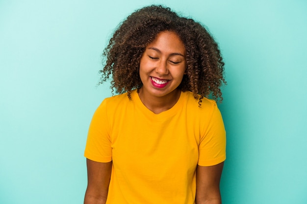 Jeune femme afro-américaine aux cheveux bouclés isolée sur fond bleu rit et ferme les yeux, se sent détendue et heureuse.