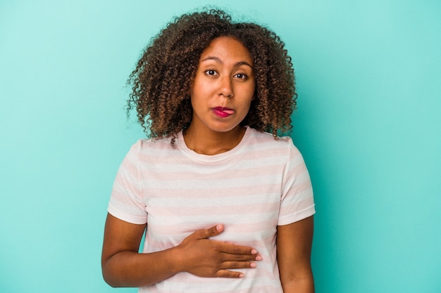 Jeune femme afro-américaine aux cheveux bouclés isolée sur fond bleu touche le ventre, sourit doucement, concept de manger et de satisfaction.