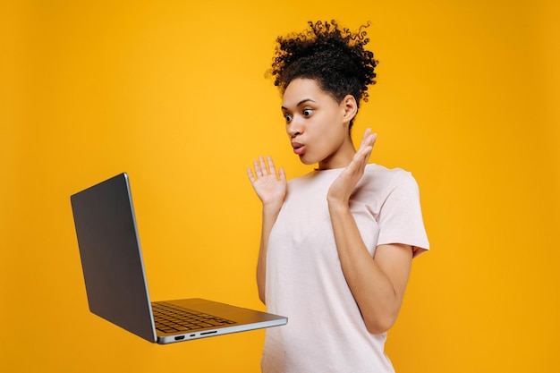 Photo une jeune femme afro-américaine aux cheveux bouclés, perplexe et choquée, regarde avec surprise l'écran de l'ordinateur portable gesticule avec ses mains a vu les kiosques à journaux inattendus sur fond orange isolé