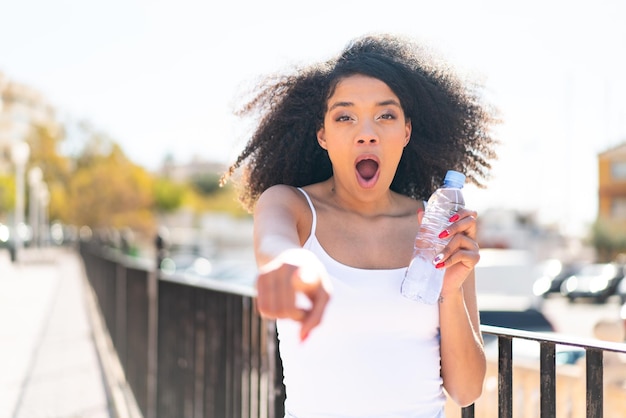 Photo jeune femme afro-américaine avec une bouteille d'eau à l'extérieur surprise et pointant vers l'avant