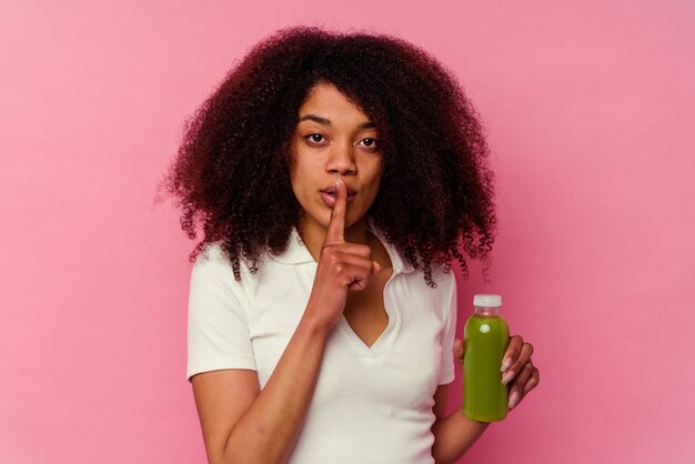 Jeune femme afro-américaine buvant un smoothie sain isolé sur fond rose en gardant un secret ou en demandant le silence.