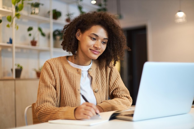 Une jeune femme afro-américaine confiante, étudiante intelligente, prend des notes tout en regardant un webinaire sur un ordinateur portable