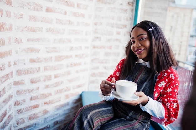 Une jeune femme afro-américaine confiante en vêtements décontractés intelligents au café avec une tasse de boisson chaude à la main