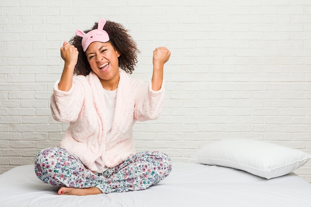 Jeune femme afro-américaine dans le lit portant pijama acclamant insouciant et excité. Concept de victoire.