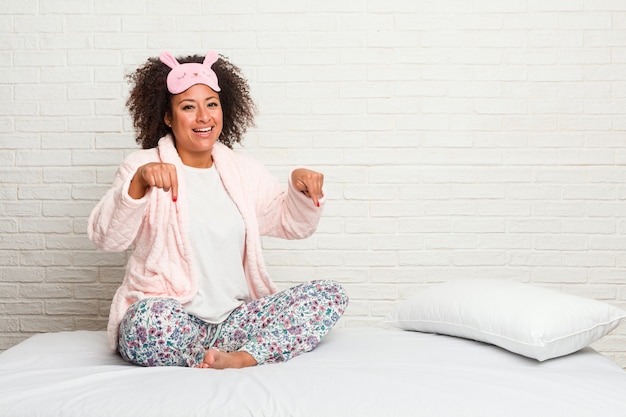 Jeune femme afro-américaine dans le lit portant pijama pointe avec les doigts, sentiment positif.