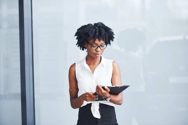 Jeune femme afro-américaine dans des verres avec bloc-notes dans les mains se trouve à l'intérieur.