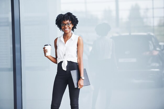 Jeune femme afro-américaine dans des verres avec bloc-notes et tasse de boisson dans les mains se trouve à l'intérieur.