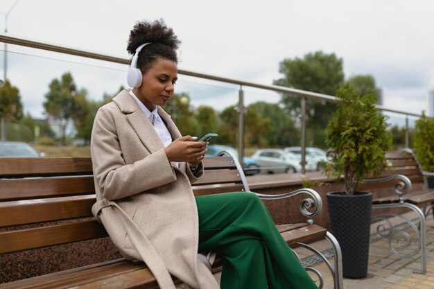 Jeune femme afro-américaine écoutant de la musique avec des écouteurs du téléphone assis sur un banc dans