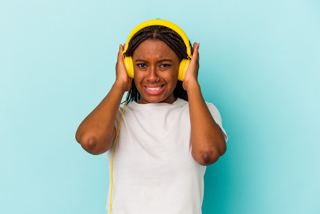 Jeune femme afro-américaine écoutant de la musique isolée sur fond bleu couvrant les oreilles avec les mains.