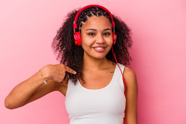 Jeune femme afro-américaine, écouter de la musique avec des écouteurs isolé sur mur rose personne pointant à la main vers un espace de copie de chemise, fier et confiant