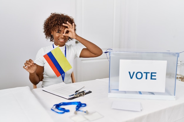 Jeune femme afro-américaine à l'élection de la campagne politique tenant le drapeau colombien souriant heureux faisant signe ok avec la main sur les yeux regardant à travers les doigts