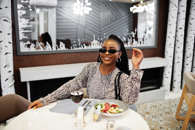 Jeune femme afro-américaine élégante, porter des lunettes de soleil noires, assis au restaurant, profiter d'une nourriture saine avec du vin.