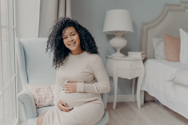 Jeune femme afro-américaine enceinte en robe blanche posant dans le salon