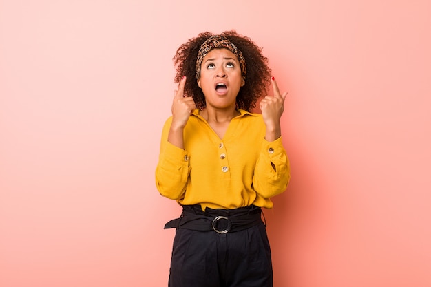 Jeune femme afro-américaine sur un fond rose pointant à l'envers avec la bouche ouverte.