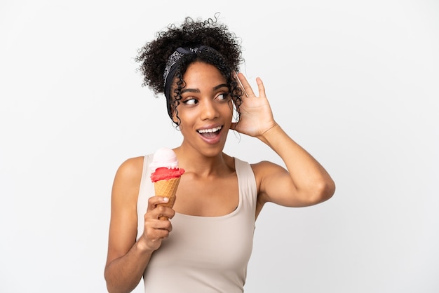 Jeune femme afro-américaine avec une glace au cornet isolée sur fond blanc écoutant quelque chose en mettant la main sur l'oreille