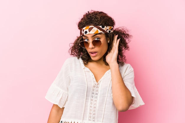 Jeune femme afro-américaine hipster isolée sur mur rose en train d'écouter un potin.