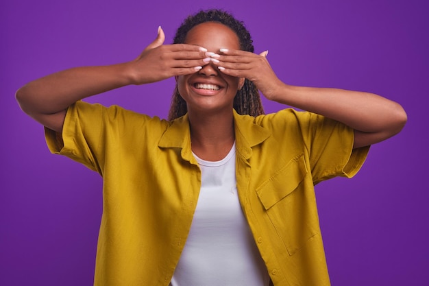 Jeune femme afro-américaine intriguée couvrant les yeux avec les mains se tient en studio