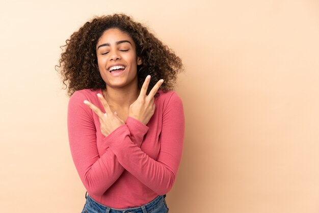 Jeune femme afro-américaine isolée sur beige souriant et montrant le signe de la victoire