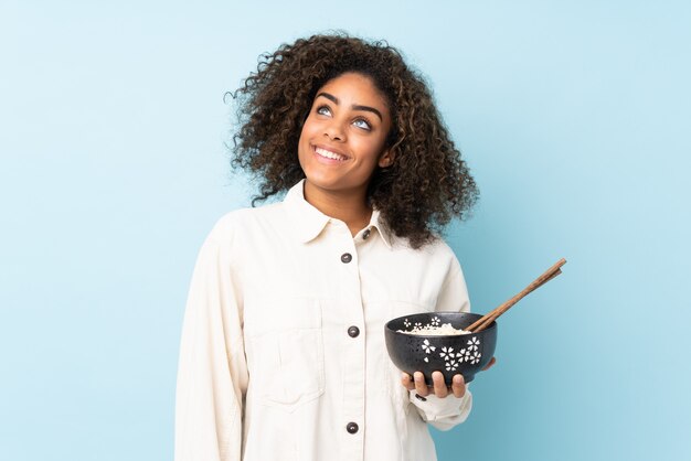 Jeune femme afro-américaine isolée sur bleu en levant tout en souriant tout en tenant un bol de nouilles avec des baguettes