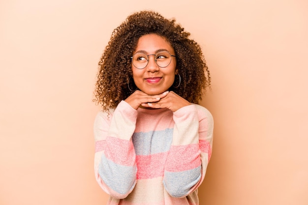 Jeune femme afro-américaine isolée sur fond beige garde les mains sous le menton regarde joyeusement de côté