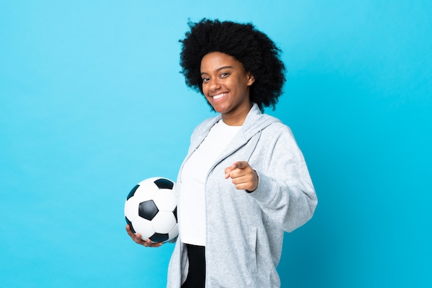 Jeune femme afro-américaine isolée sur fond bleu avec un ballon de soccer et pointant vers l'avant