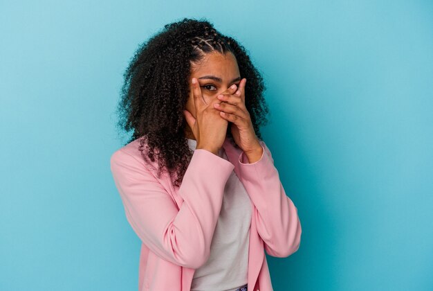 Jeune femme afro-américaine isolée sur fond bleu clignote à travers les doigts effrayés et nerveux.