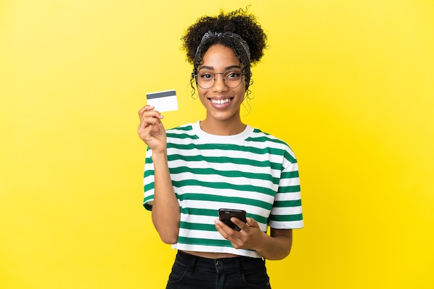 Jeune femme afro-américaine isolée sur fond jaune achetant avec le mobile avec une carte de crédit