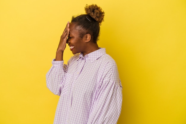 Jeune femme afro-américaine isolée sur fond jaune clignote à l'appareil photo à travers les doigts, gêné couvrant le visage.