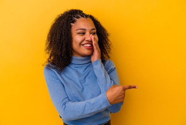 Jeune femme afro-américaine isolée sur fond jaune disant un potin, pointant vers le côté rapportant quelque chose.