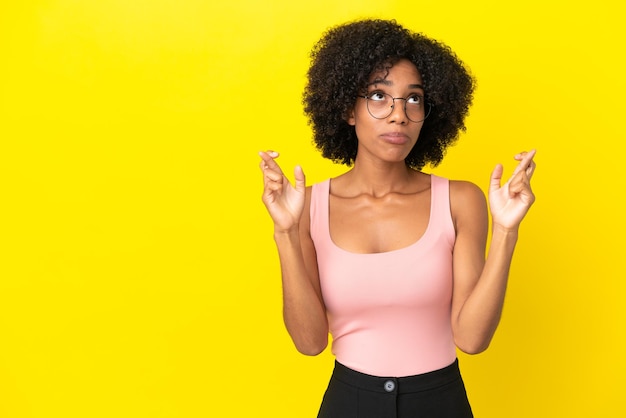 Jeune femme afro-américaine isolée sur fond jaune avec les doigts croisés et souhaitant le meilleur