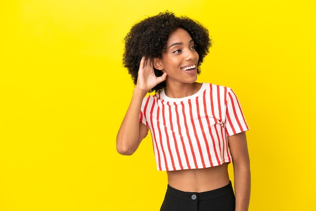 Jeune femme afro-américaine isolée sur fond jaune écoutant quelque chose en mettant la main sur l'oreille