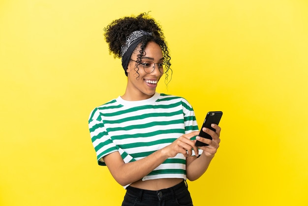 Jeune femme afro-américaine isolée sur fond jaune envoyant un message ou un e-mail avec le mobile