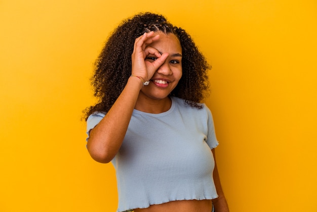 Jeune femme afro-américaine isolée sur fond jaune excité en gardant le geste ok sur les yeux.