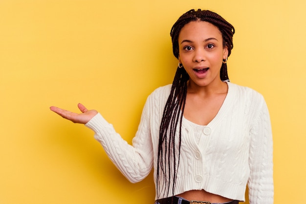 Jeune femme afro-américaine isolée sur fond jaune impressionné tenant un espace de copie sur la paume.