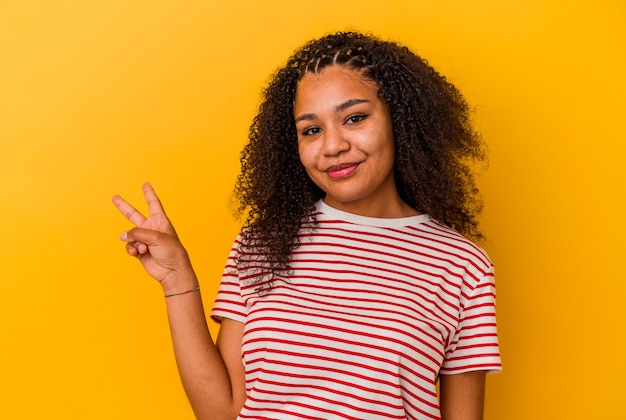 Jeune femme afro-américaine isolée sur fond jaune montrant le signe de la victoire et souriant largement.