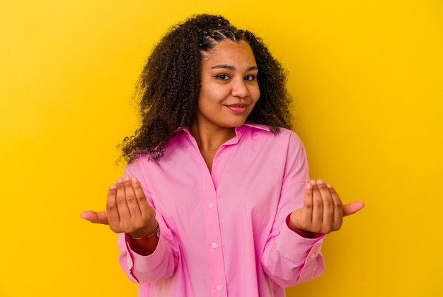 Photo jeune femme afro-américaine isolée sur fond jaune pointant avec le doigt sur vous comme si vous invitiez à vous rapprocher.