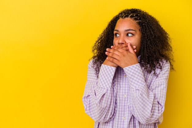 Jeune femme afro-américaine isolée sur fond jaune réfléchie à la recherche d'un espace de copie couvrant la bouche avec la main.