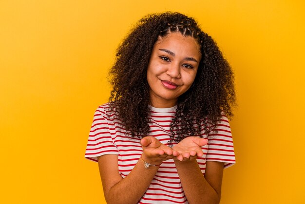 Jeune femme afro-américaine isolée sur fond jaune se pliant les lèvres et tenant les paumes pour envoyer l'air baiser.