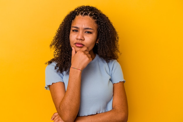 Jeune femme afro-américaine isolée sur fond jaune suspect, incertain, vous examine.