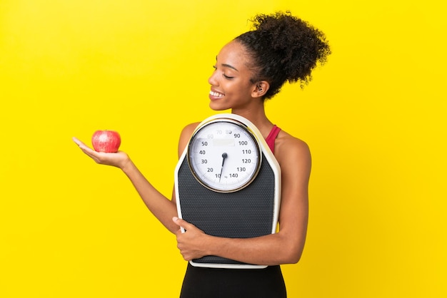 Jeune femme afro-américaine isolée sur fond jaune tenant une balance tout en regardant une pomme