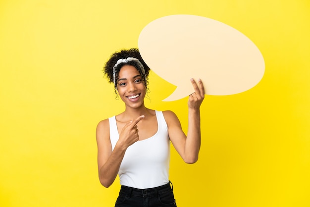 Jeune femme afro-américaine isolée sur fond jaune tenant une bulle de dialogue vide et la pointant