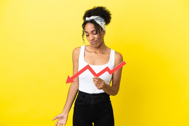 Jeune femme afro-américaine isolée sur fond jaune tenant une flèche vers le bas et avec une expression triste