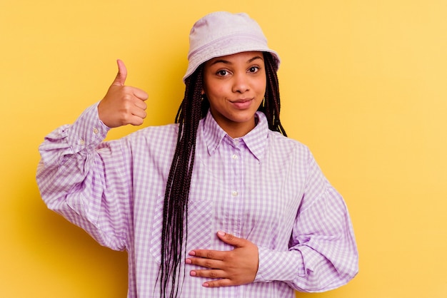 Jeune femme afro-américaine isolée sur fond jaune touche le ventre, sourit doucement, concept de manger et de satisfaction.