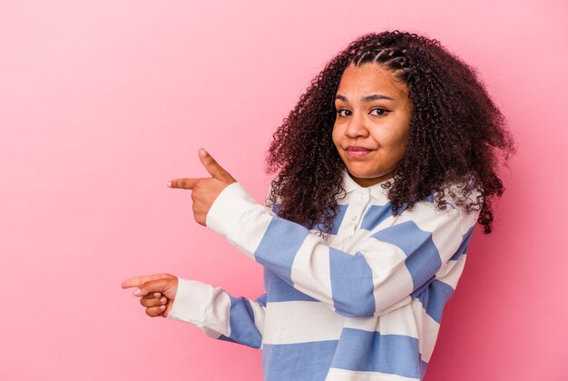 Jeune femme afro-américaine isolée sur fond rose choquée pointant avec l'index vers un espace de copie.