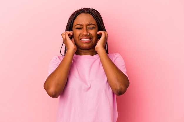 Jeune femme afro-américaine isolée sur fond rose couvrant les oreilles avec les mains.