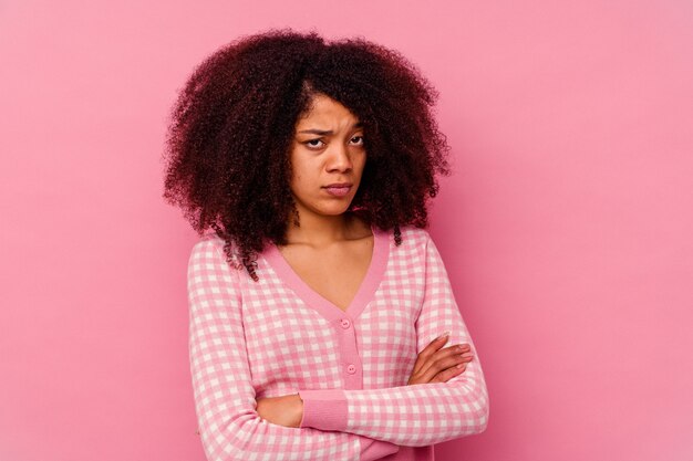 Jeune femme afro-américaine isolée sur fond rose fronçant le visage de mécontentement, garde les bras croisés.