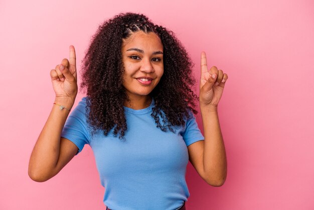 Jeune femme afro-américaine isolée sur fond rose indique avec les deux doigts avant montrant un espace vide.