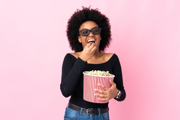 Jeune femme afro-américaine isolée sur fond rose avec des lunettes 3D et tenant un grand seau de pop-corn