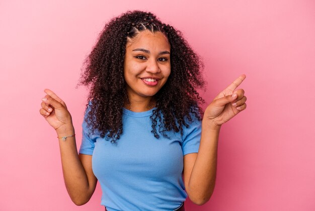 Jeune femme afro-américaine isolée sur fond rose pointant vers différents espaces de copie, en choisissant l'un d'entre eux, montrant avec le doigt.